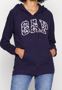 Felpa con zip di colore blu navy con logo "GAP" in stampa floreale, dotata di cappuccio con lacci, maniche lunghe e tasche anteriori. Tessuto morbido in misto cotone.