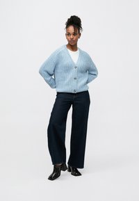 Cardigã azul-claro de malha com decote em V, fecho de botões e padrão texturizado; combinado com jeans escuros de perna larga e sapatos pretos com salto.