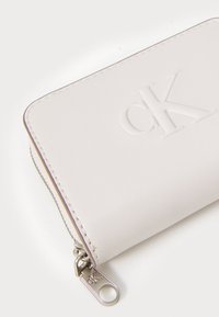 Portafoglio in pelle bianca con logo "CK" in rilievo, chiusura con zip e dettagli in metallo argentato. Texture liscia con bordi cuciti e angoli arrotondati.
