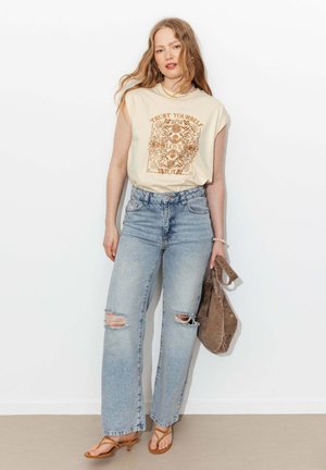 Jeune femme aux longs cheveux porte un haut sans manches crème à imprimé floral avec le texte « Trust Yourself », jean bleu clair déchiré, sandales et tient un sac en daim.