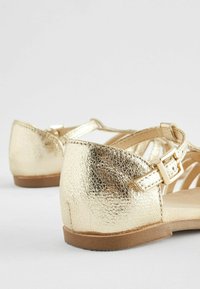 Guldmetalliska sandaler med en texturerad yta, öppen tå-design och en ankelrem med en metallspänne, vilande på en ljusbrun sula.