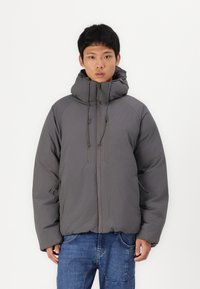 Sivi puffer jacket s visokim ovratnikom i podesivim vezicama. Ima zatvarač sprijeda i dva prednja džepa. Ležeran kroj s mat završnom obradom.