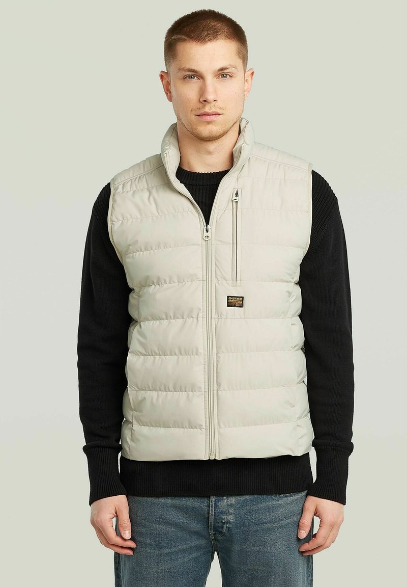 Gilet imbottito beige chiaro con zip frontale, colletto alto e tasca sul petto; indossato sopra un maglione nero a maglia, caratterizzato da una vestibilità rilassata.