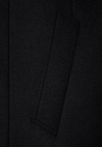 Czarna wełniana tkanina płaszcza o miękkiej teksturze z prostokątnym detalem kieszeni. Widoczne szwy wzdłuż szwów podkreślają usztywnioną konstrukcję.