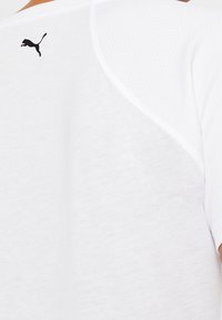 Valkoinen urheilupaita, jossa on teksturoitu kangas, musta Puma-logo yläselässä ja sileät lyhyet hihat.