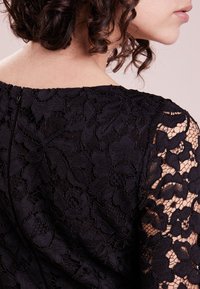 Schwarzspitzenoberteil mit aufwendigen Blumenmustern, transparenten Ärmeln und einem dezenten Reißverschluss auf der Rückseite, der den strukturierten Stoff hervorhebt.