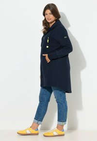 Námořnicky modré mikinové šaty s texturovanou látkou, předními kapsami a neonovými šňůrkami. Kombinované s světle modrými džíny a žlutými teniskami s detaily.