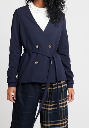 Kobieta w granatowej dwurzędowej marynarce z paskiem, na białej bluzce z falbanami oraz szerokich spodniach z mieszanymi wzorami w kratę.
