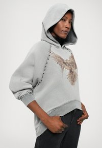 Gray hoodie with a large eagle graphic, drawstring hood, and unique beaded detailing along the seams. Soft fabric with ribbed cuffs.