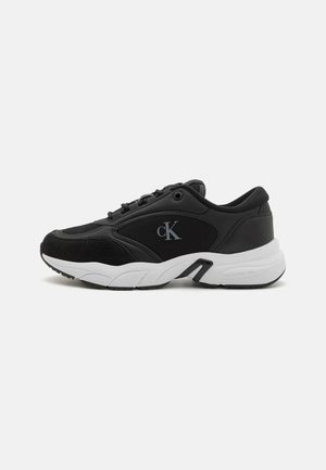 Černé sportovní boty se kombinací hladké kůže a semiše, s robustní bílou podrážkou a výrazným logem na boku.