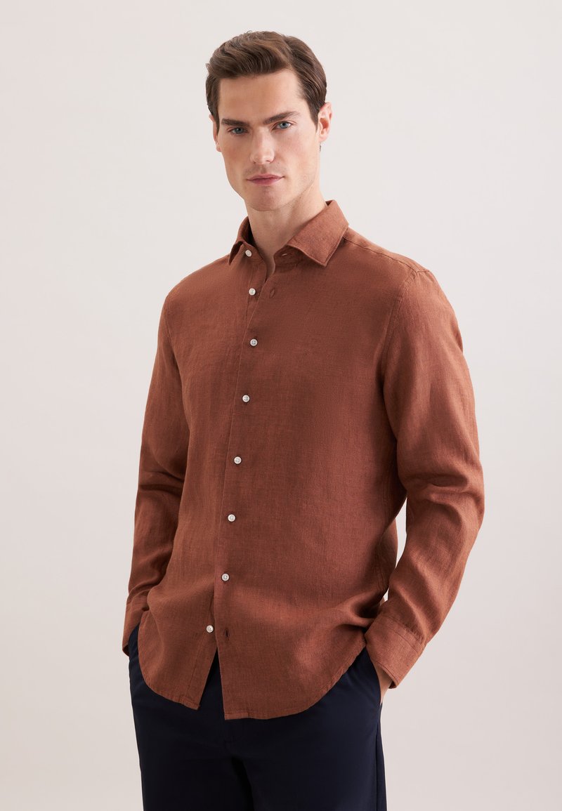Homme portant une chemise marron à manches longues boutonnée avec col et un pantalon bleu marine foncé, debout les mains dans les poches devant un fond neutre.
