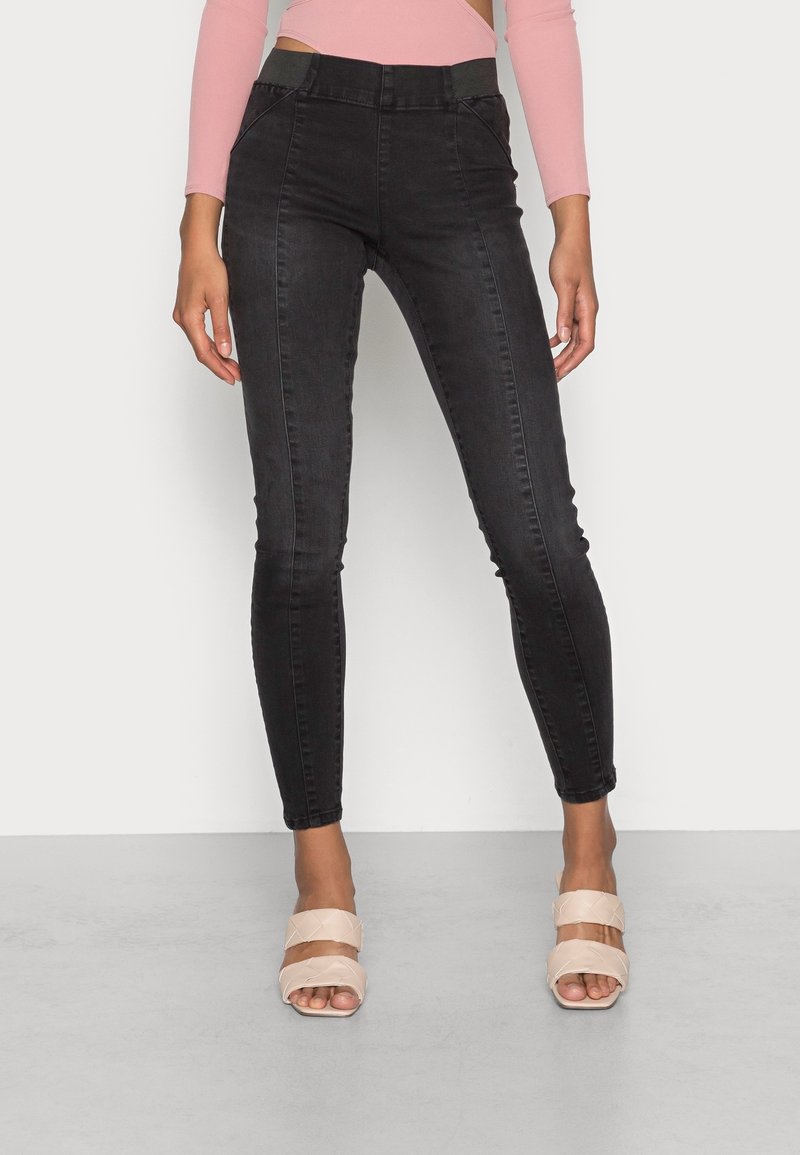 Femme portant un jean skinny noir taille haute associé à un crop top à manches longues rose clair et des sandales beiges à talons ouverts.