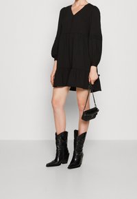 Abito lungo nero con maniche lunghe, scollo a V, dettagli con bottoni e orlo a strati. Abbinato a stivali cowboy neri con motivo e una piccola borsa nera.