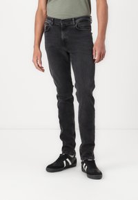 Uske slim-fit crne traperice s glatkom teksturom, s pet džepova i patentnim zatvaračem. Nosite ih s crnim tenisicama i zelenom majicom.