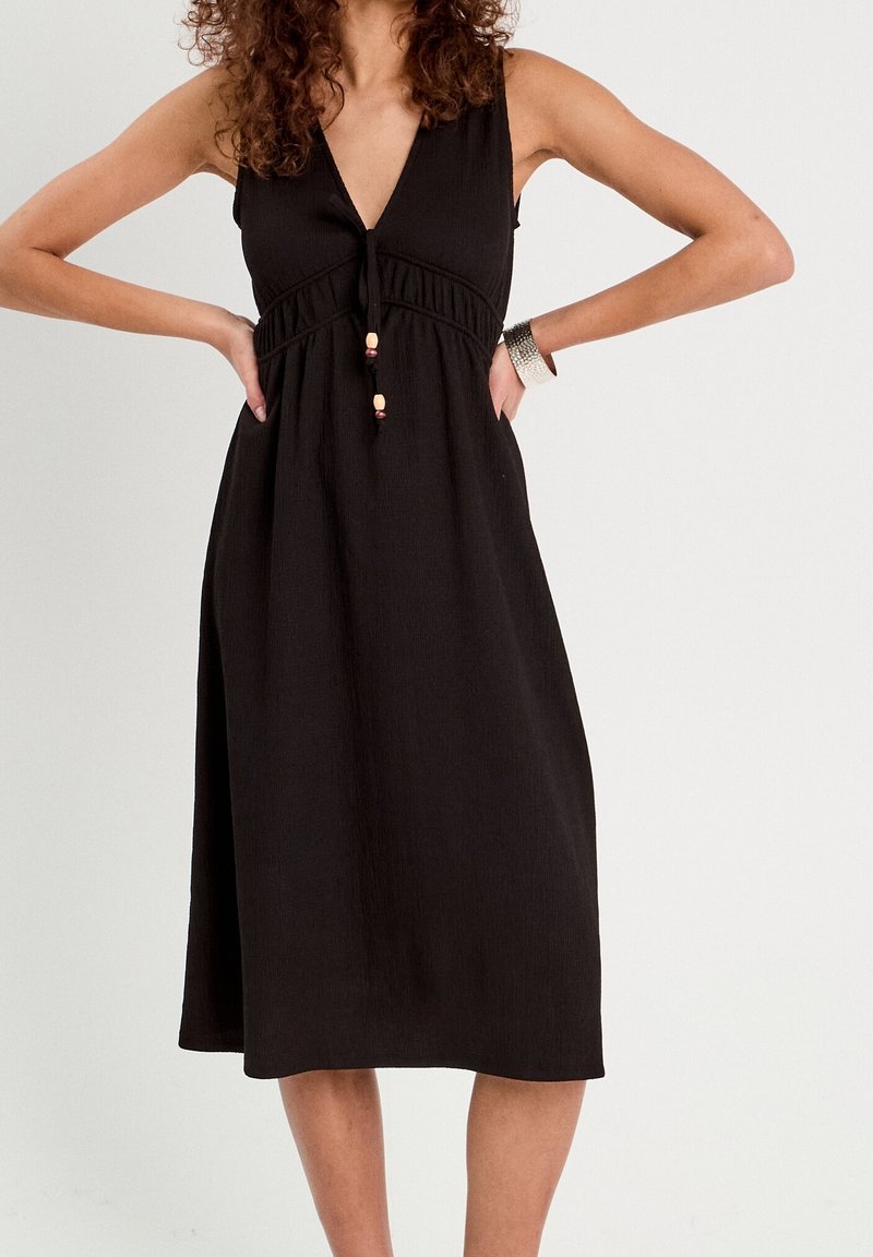 Femme portant une robe midi noire sans manches avec un col en V et un détail noué à l'avant, les mains sur les hanches, sur un fond clair uni.