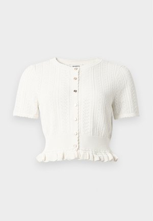 Femme portant un haut blanc à manches courtes avec boutons et une jupe noire asymétrique avec des détails en œillets dorés ainsi que des ballerines noires.