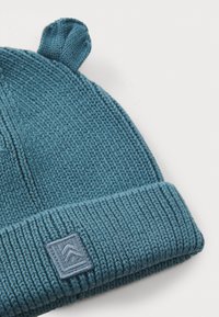 Blauwe gebreide beanie met een omgeslagen ribgebreide rand, voorzien van twee kleine oortjes en een vierkante logo patch op de rand.