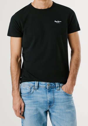 Schwarzes Baumwoll-T-Shirt mit Rundhalsausschnitt und kurzen Ärmeln, mit einem weißen Logo auf der linken Brust. Kombiniert mit hellblauen Jeans.