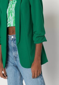 Personne portant un blazer vert aux manches retroussées sur un crop top vert à motifs blancs et un jean taille haute bleu clair.
