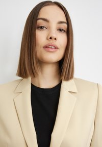 Beige blazer with sharp lapels over a black sweater. Smooth fabric, fitted design, and straight cut, with a clean neckline and no visible embellishments.