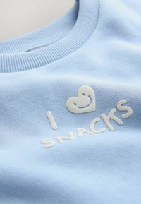 Sudadera azul claro con cuello acanalado. Presenta texto blanco, "I ❤️ SNACKS," y un gráfico de corazón sonriente en un diseño liso y en relieve.