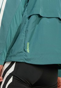 Tirkizna lagana jakna s teksturiranom površinom, koja ima ADIZERO logo u neon žutoj boji, slojevita preko crnih hlača s bijelim prugama.