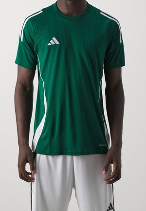 Mężczyzna ubrany w ciemnozieloną sportową koszulkę Adidas z białymi paskami oraz białe spodenki Adidas, stojący z rękami swobodnie opuszczonymi wzdłuż ciała.