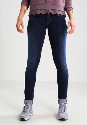Jeans ajustados en denim azul oscuro con una textura suave. Llevados con una camiseta gris y zapatillas grises a juego. Con el dobladillo enrollado en el tobillo.