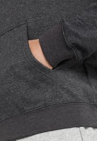 En hand inskjuten i framlommen på en mörkgrå sweatshirt med ribbad mudd och nederkant, som visar den mjuka tygtexturen.