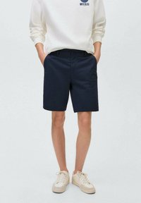 Marinblå bomullsshorts med rak passform, utrustade med bältesöglor och sidofickor, parat med en krämfärgad sweatshirt och ljusa sneakers.