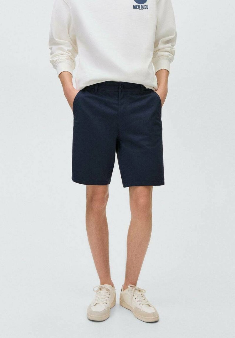 Marinblå bomullsshorts med rak passform, utrustade med bältesöglor och sidofickor, parat med en krämfärgad sweatshirt och ljusa sneakers.