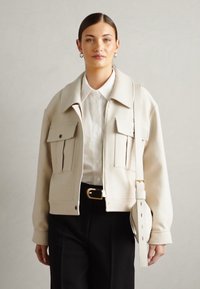 Beige kurz geschnittene Jacke aus glattem Material, ausgestattet mit zwei großen Fronttaschen, einem klassischen Kragen und einem verstellbaren Riemen.