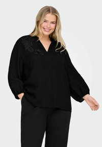 Blusa negra texturizada con escote en V y mangas abullonadas, que presenta bordados florales en un hombro. Combinada con pantalones negros a juego.