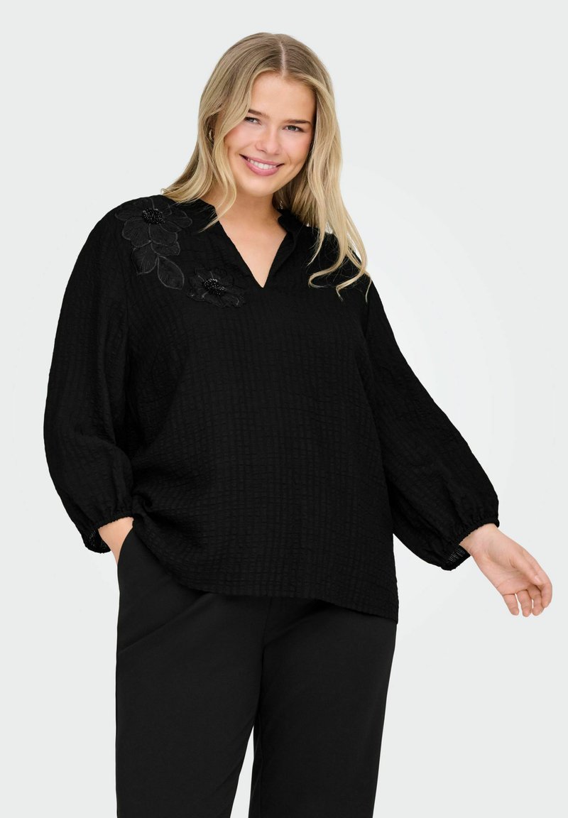 Blusa negra texturizada con escote en V y mangas abullonadas, que presenta bordados florales en un hombro. Combinada con pantalones negros a juego.