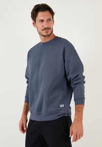 Felpa blu scuro realizzata in tessuto morbido, con collo a giro, maniche lunghe e un'etichetta sul lato sinistro in basso.