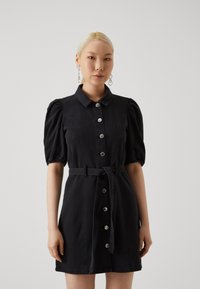 Robe en denim noir avec de courtes manches bouffantes, boutonnée sur le devant et ceinturée à la taille. Présente des boutons en argent et des coutures structurées.