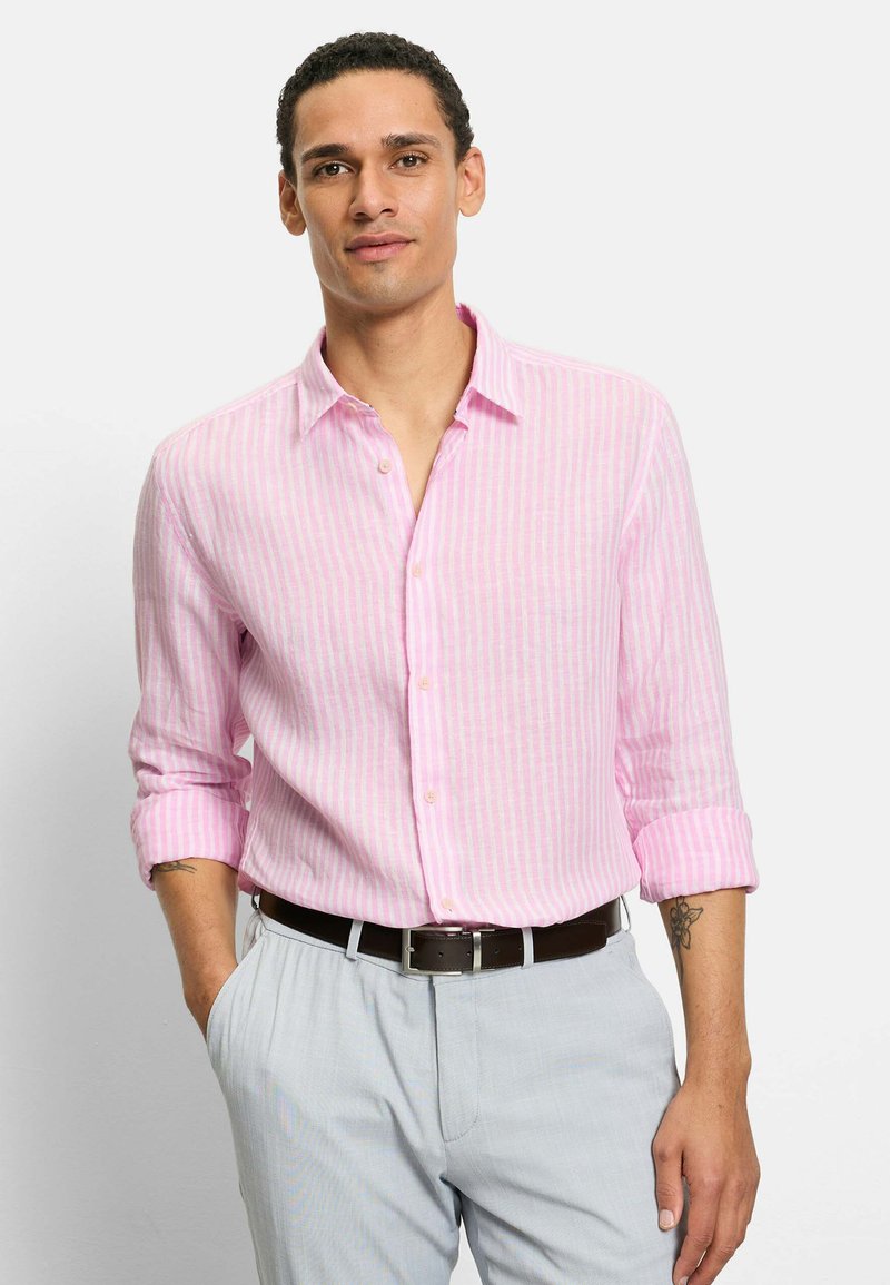 Mann mit kurzen lockigen Haaren, der ein rosa-weiß gestreiftes Hemd mit langen Ärmeln und hellgraue Hosen trägt, steht mit einer Hand in der Tasche.