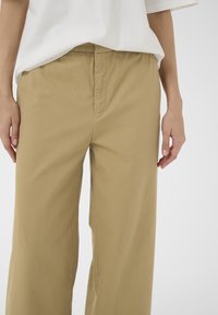 Partie centrale d'une personne portant un pantalon beige ample et une chemise blanche, les mains détendues le long du corps, sur un fond clair uni.