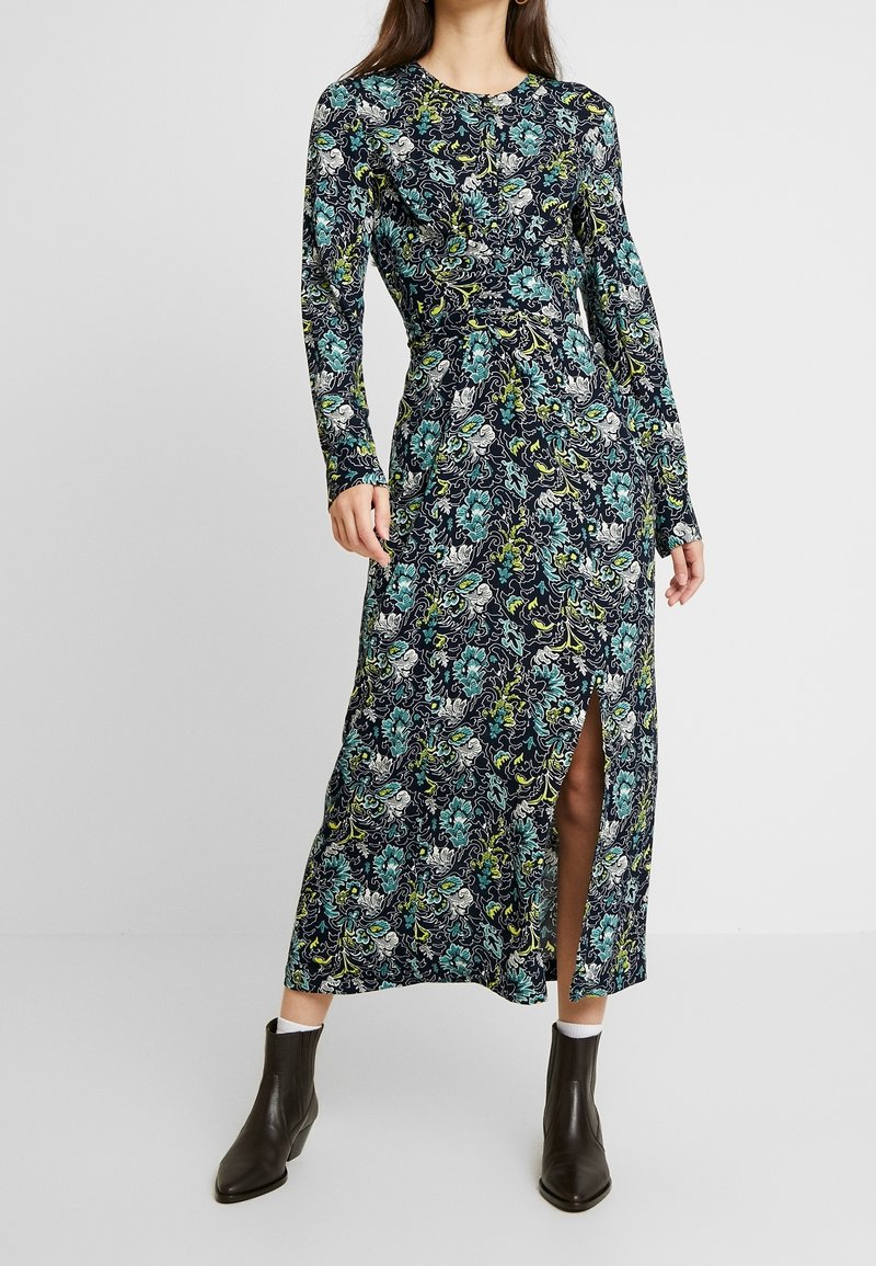 Långärmad blommig klänning med en mörk marinblå bakgrund och intrikata mönster i grönt och blått samt en hög slits till låret. Tillverkad av mjukt tyg.