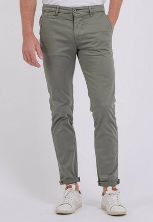Homme portant un pantalon slim olive retroussé et des baskets blanches, debout devant un fond blanc.