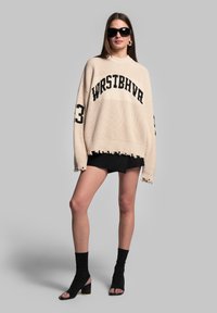 Beige gestrickter Pullover mit Rippstruktur, schwarzer Schrift und ausgefransten Kanten, kombiniert mit schwarzen Shorts und offenen Schuhe mit Blockabsatz.