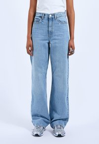 Světle modré džíny s vysokým pasem a širokým střihem, vyrobené z denimu. Mají klasické kapsy a zipový kloup. Jsou v kombinaci se šedými teniskami.