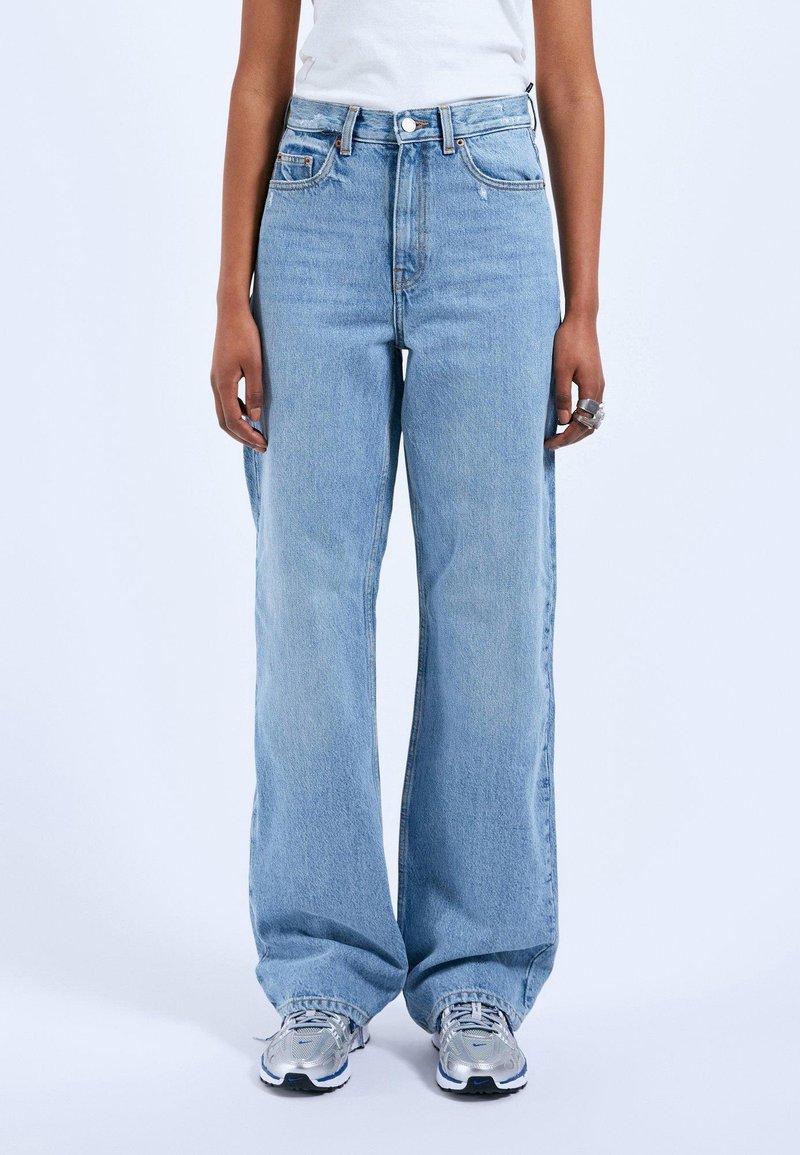 Světle modré džíny s vysokým pasem a širokým střihem, vyrobené z denimu. Mají klasické kapsy a zipový kloup. Jsou v kombinaci se šedými teniskami.