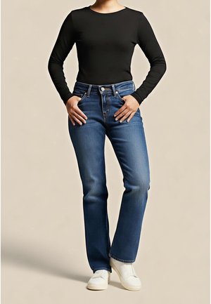 Person iført en tætsiddende sort langærmet top, blå jeans med lige ben, hvide slip-on sko, stående med hænderne i forlommerne.