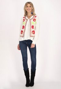 Weiße Bluse mit roter und grüner Floralstickerei auf der Vorderseite, kombiniert mit blauen, engen Jeans und schwarzen Wildleder-Cowboy-Stiefeln.