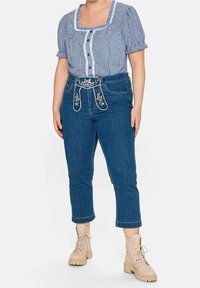 Blå rutmönstrad blus med puffärmar, knäppning fram, ihop med åtsittande mörkblå jeans med ett broderat bälte och beige kängor.