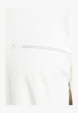 Aktivna majica v off-white barvi, izdelana iz mehkega materiala, z rahlo ukrivljenim šivom in minimalnimi teksturiranimi detajli, ter natisnjenim besedilom ob strani.