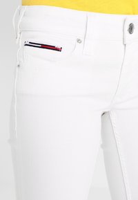Bílé denimové džíny s přiléhavým střihem, s předními kapsami a malým barevným pruhovaným akcentem na levé kapse.