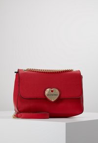 Borsa a mano in pelle rossa con finitura testurizzata, hardware a forma di cuore in oro, pattina pieghevole e tracolla a catena. Forma compatta con tracolla staccabile.
