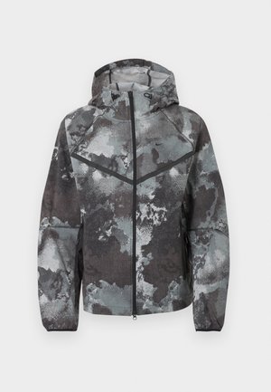 Heren jas met capuchon en volledige rits aan de voorkant, camouflagepatroon in grijstinten, zwarte chevron detail en ritszakken aan de zijkanten.